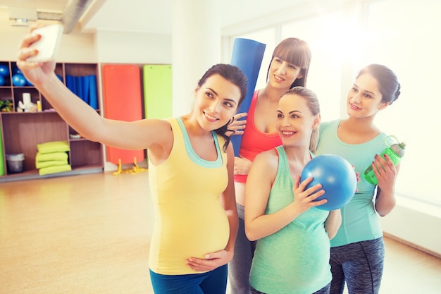 Foto gravidanza, sport, fitness, persone e concetto di stile di vita sano - gruppo di donne incinte felici con cose sportive che prendono selfie con lo smartphone in palestra