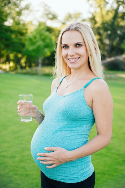 Беременность, спорт и здоровый образ жизни - счастливая беременная женщина пьет воду из стакана в парке в солнечный летний день