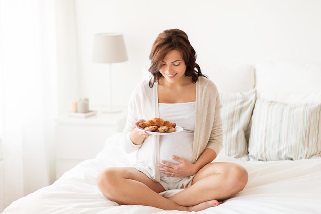 pregnancy, unhealthy eating, food and people concept - happy pregnant woman with croissant buns at home