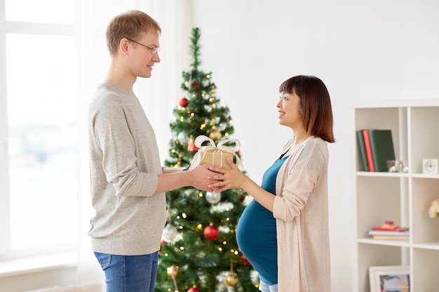 妊娠,冬の休日,人々のコンセプト - 幸せな夫が妊娠中の妻にクリスマスプレゼントを家で与える