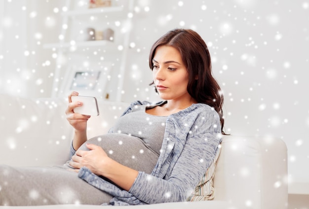 pregnancy, winter, technology, people and expectation concept - sad pregnant woman with smartphone at home over snow