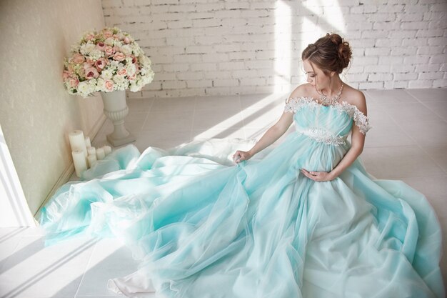 Беременность, женщина сидит на полу в роскошном платье и держит руки за живот.