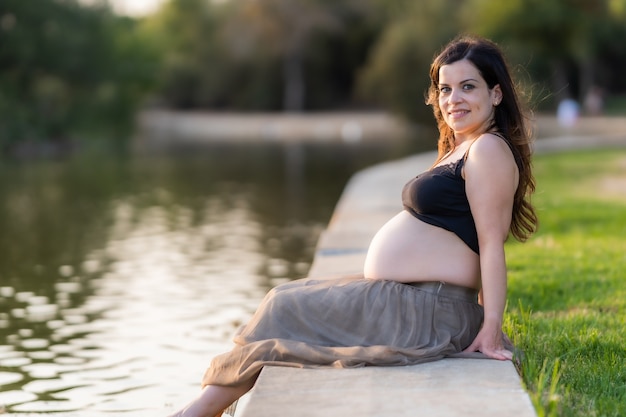 Pregnant adult Latina woman sitting on the edge of a river in a park with a happy expression wearing comfortable summer clothes during sunset