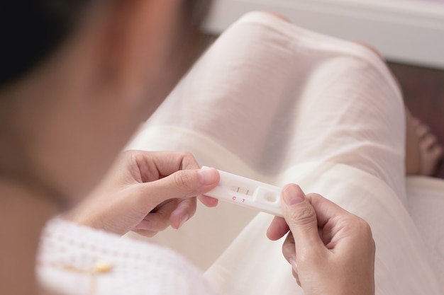 Donna incinta dell'asia in vestito bianco che esamina la striscia di test di gravidanza