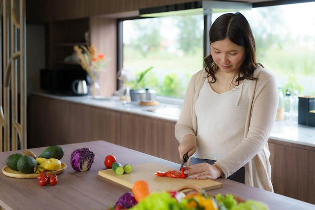 妊娠中のアジア人女性が新鮮な緑のサラダのためにトマトを切る女性が美味しい有機的な夕食を準備する