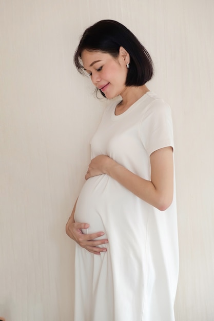 A pregnant Asian woman wearing casual clothes is standing and looking her belly