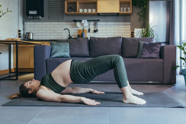 Беременная красивая женщина занимается спортом дома сидит и лежит на спортивном коврике в спортивной форме делает упражнения довольна