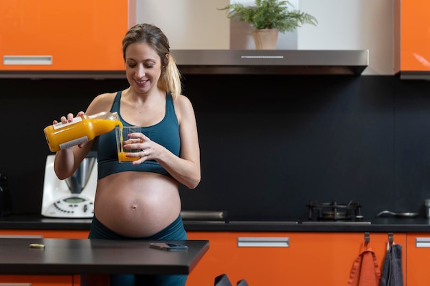 Pregnant blonde woman drinking an orange juice in the kitchen She is wearing sport clothes