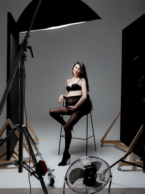 Pregnant brunette girl in pantyhose bra and shoes on a high chair in a photo studio