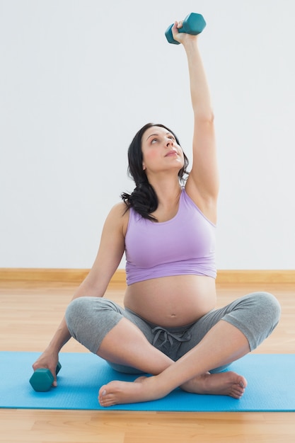 Pregnant brunette sitting on exercise mat lifting hand weight