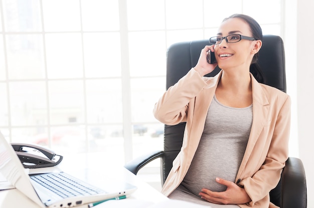 Pregnant business lady at work. Cheerful pregnant businesswoman talking on the phone while sitting at her working place in office