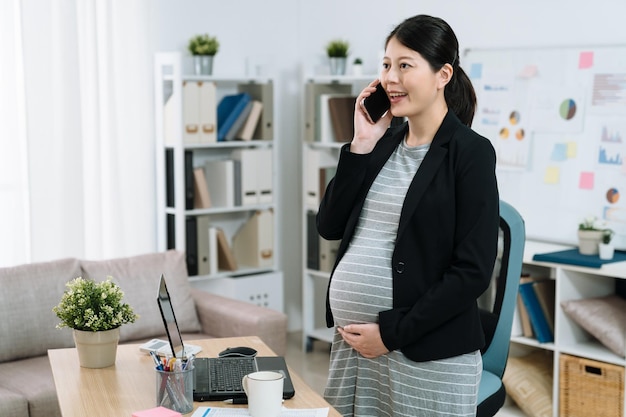 Donna d'affari incinta che parla al cellulare mentre è in piedi vicino alla scrivania in un ufficio luminoso e accogliente. giovane futura mamma lavoratrice in giacca e cravatta con conversazione sul cellulare e mano che tiene grande pancia all'interno