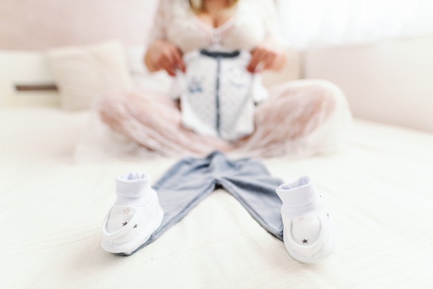 Pregnant Caucasian woman holding baby's clothes wile sitting on the bed with legs crossed in bedroom. Selective focus technique.