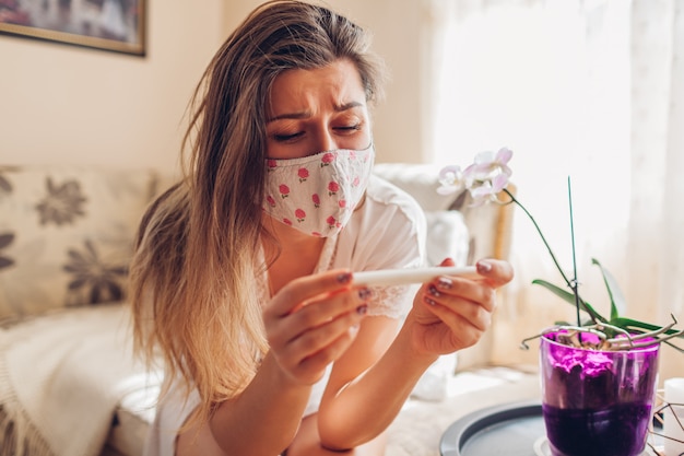 Беременность во время коронавируса ковид-19. Больная женщина в маске проверяя тест на беременность дома.
