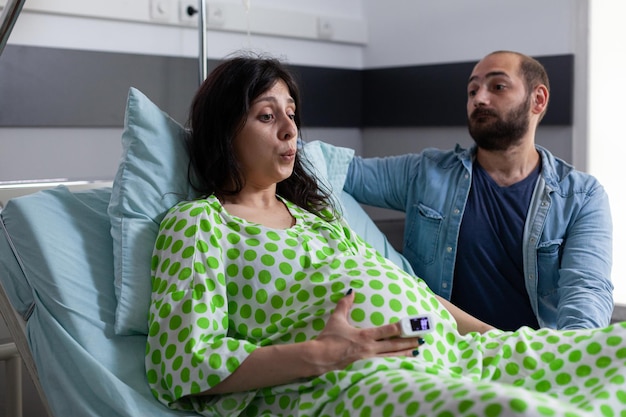 Coppia incinta che respira mentre aspetta un bambino in una struttura medica nel reparto ospedaliero. genitori caucasici che si preparano per il parto e la genitorialità mentre la giovane donna è seduta a letto