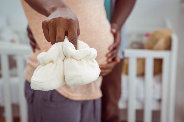 Pregnant couple holding white baby shoes near cradle