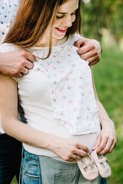 Pregnant couple put baby shoes and clothes on stomach, waiting baby.