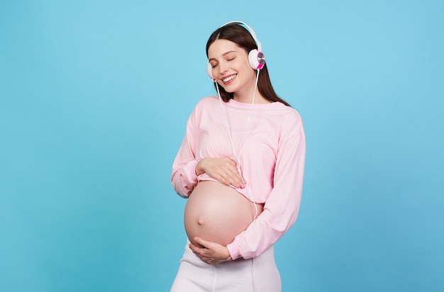 임신한 귀여운 젊은 여성은 파란색 배경에 헤드폰으로 음악을 듣습니다.