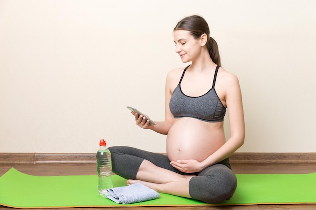 Pregnant female holding smartphone while sitting on exercise yoga mat at home at coronavirus time