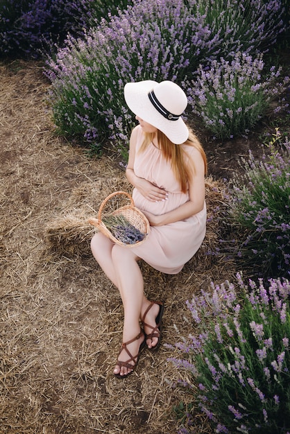 Pregnant girl blonde in a beige dress and straw hat. Lavender field. In anticipation of a child. The idea of a photo shoot. Walk at sunset. Future mom. Basket of flowers.