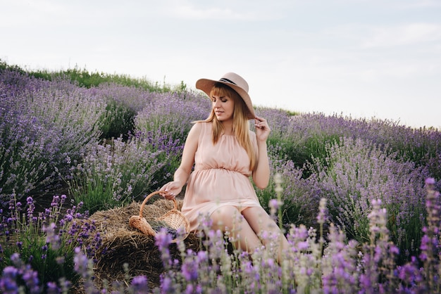 Pregnant girl blonde in a beige dress and straw hat. Lavender field. In anticipation of a child. The idea of a photo shoot. Walk at sunset. Future mom. Basket of flowers.