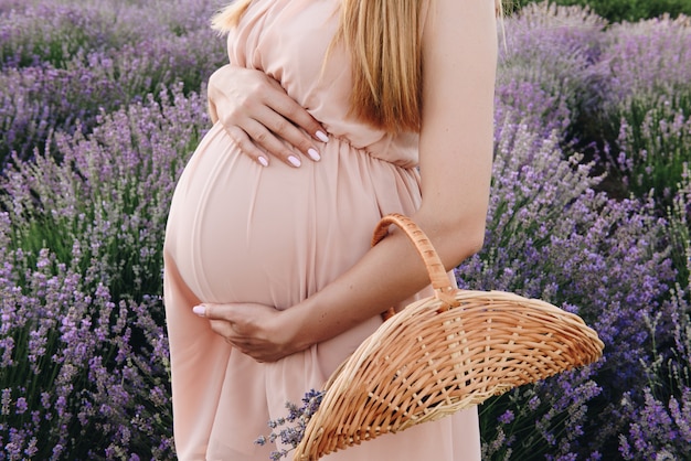 妊娠中の女の子金髪のベージュのドレスと麦わら帽子。ラベンダー畑。子供を見越して。写真撮影のアイデア。夕暮れを歩く。将来のお母さん。花のバスケット。