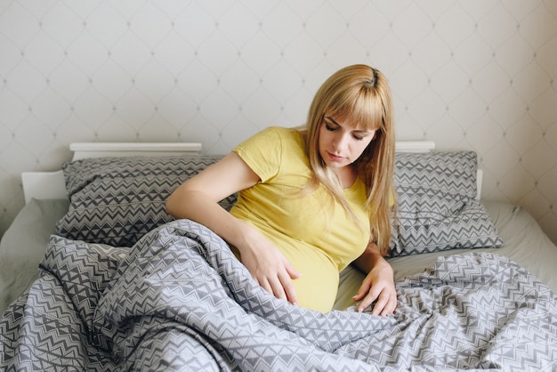 Pregnant girl blonde in a yellow T-shirt at home. Waiting for a miracle. Pregnancy. Love and hope. Light room. Lies in bed in the bedroom. Gray bedding.