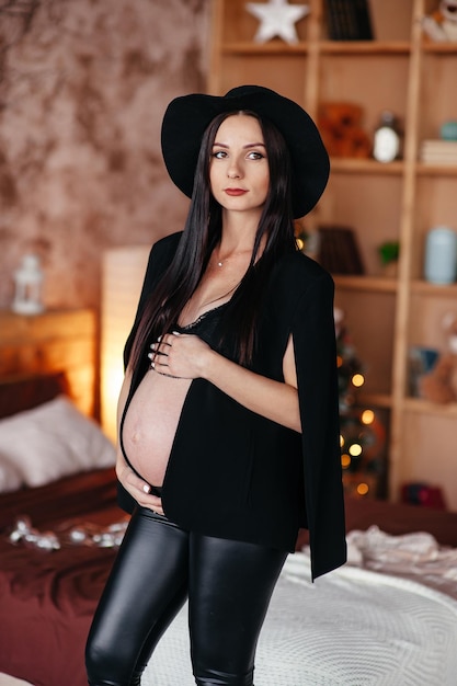 Pregnant girl dressed in black dress and hat holds herself tummy