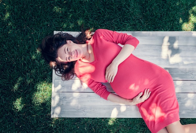 Pregnant girl dressed in pink dress, lying on a grass and holds herself tummy