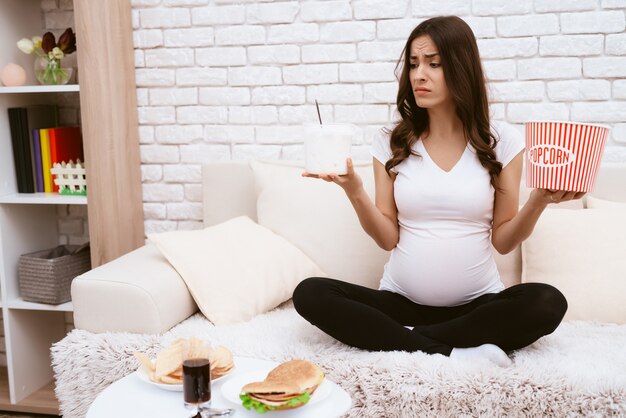 A pregnant girl eats a lot of popcorn on the couch.