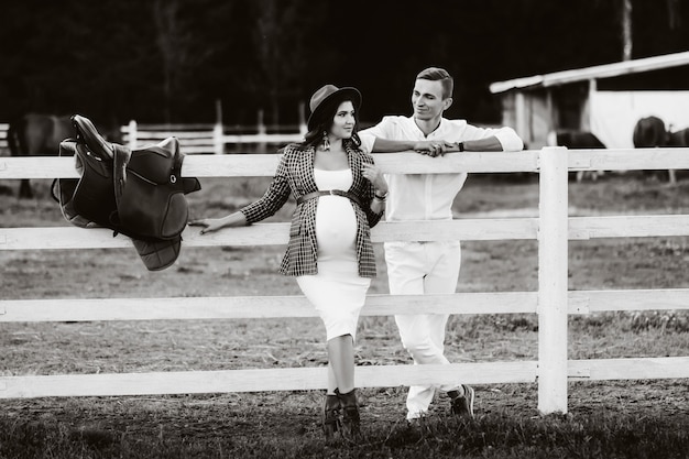 A pregnant girl in a hat and her husband in white clothes stand next to the horse corral.a stylish couple waiting for a child stand on the street near the horse corral. black and white photo.