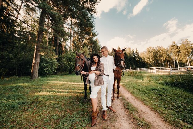 A pregnant girl in a hat and her husband in white clothes stand next to horses in the forest in nature