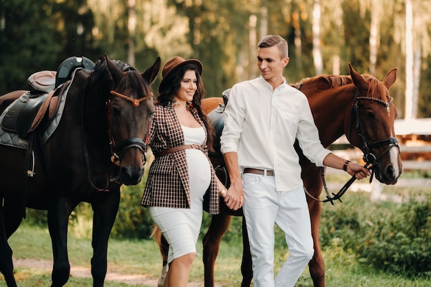 A pregnant girl in a hat and her husband in white clothes stand next to horses in the forest in nature