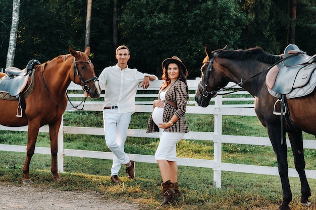 A pregnant girl in a hat and a man in white clothes stand next to horses near a white fence.Stylish pregnant woman with a man with horses.Married couple.