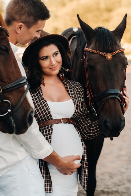 A pregnant girl in a hat and a man in white clothes stand next to horses near a white fence.Stylish pregnant woman with a man with horses.Married couple.