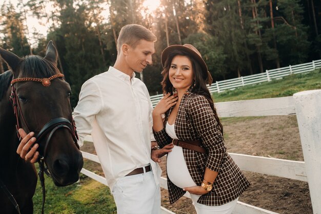 A pregnant girl in a hat and a man in white clothes stand next to horses near a white fence.Stylish pregnant woman with a man with horses.Married couple.