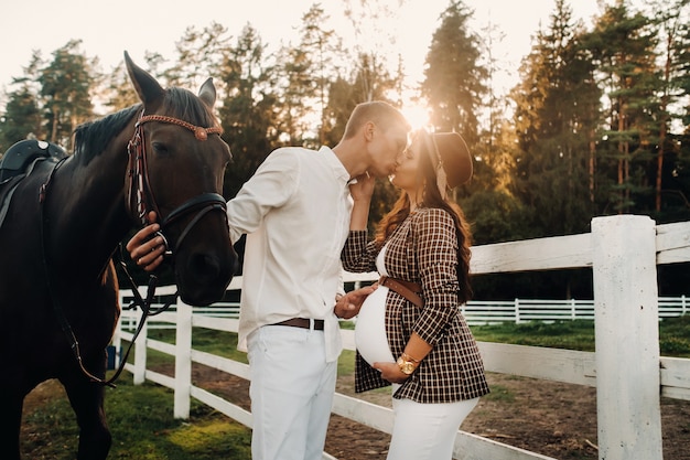 A pregnant girl in a hat and a man in white clothes stand next to horses at a White fence.