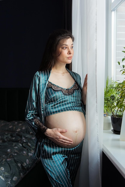 Pregnant girl in pajamas standing by the window