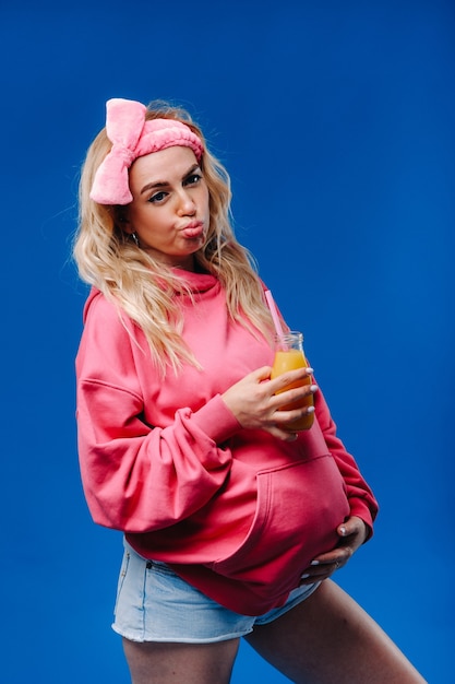 Pregnant girl in pink clothes with a bottle of juice on a blue background