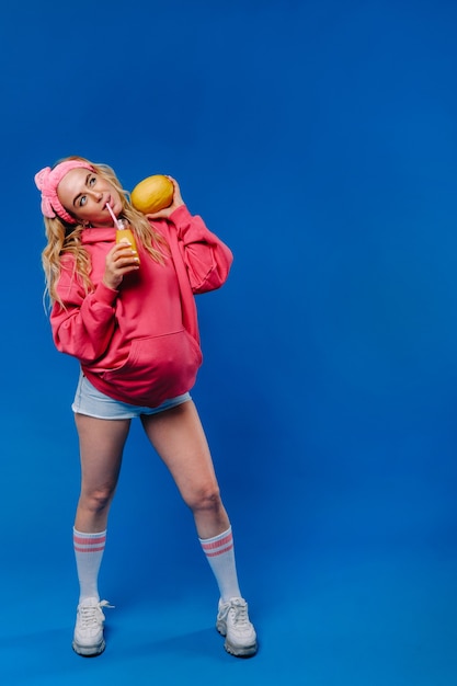 Pregnant girl in pink clothes with a bottle of juice and a melon on a blue background