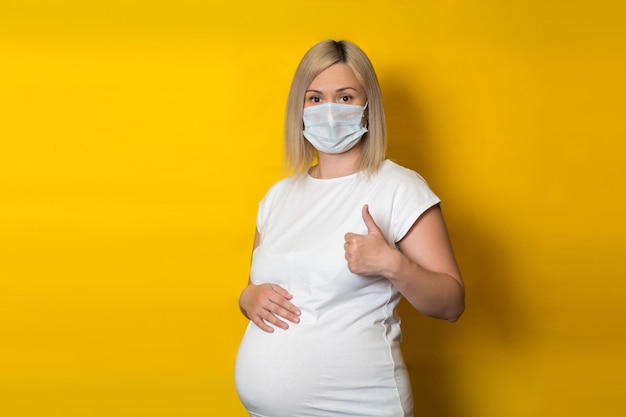 A pregnant girl in a protective mask makes a thumbs-up gesture and with her hand on her stomach. healthcare concept