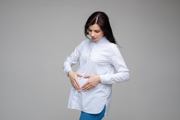 妊娠中の女の子は、赤ちゃんを待っている腹にハートの形のジェスチャーを示しています。マタニティの概念。妊娠中の妊婦