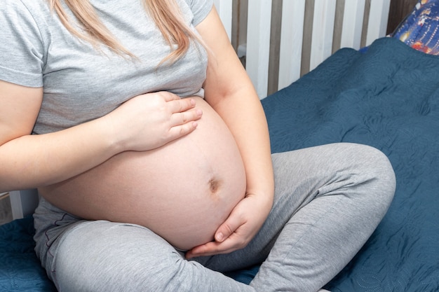 A pregnant girl sits on the bed at home and holds her hands on her stomach