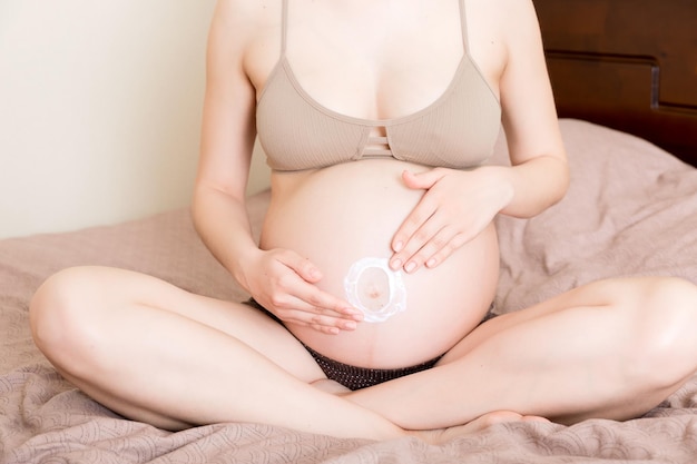 a pregnant girl sits at home on the bed and smears an antistretch mark cream on her stomach Pregnancy motherhood preparation and expectation concept