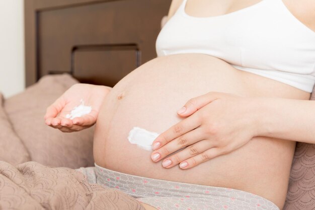 a pregnant girl sits at home on the bed and smears an antistretch mark cream on her stomach Pregnancy motherhood preparation and expectation concept