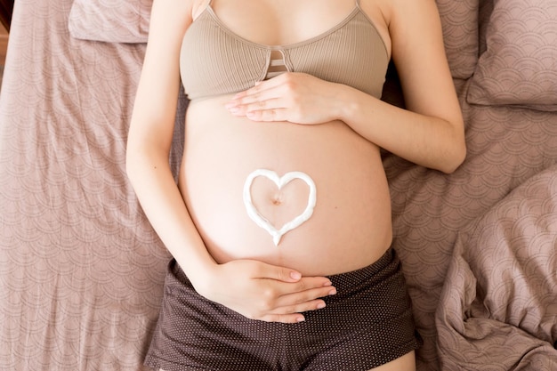 임신한 소녀는 집에 침대에 앉아 배에 항스트레치 마크 크림을 바르고 있습니다. 임신 모성 준비 및 기대 개념