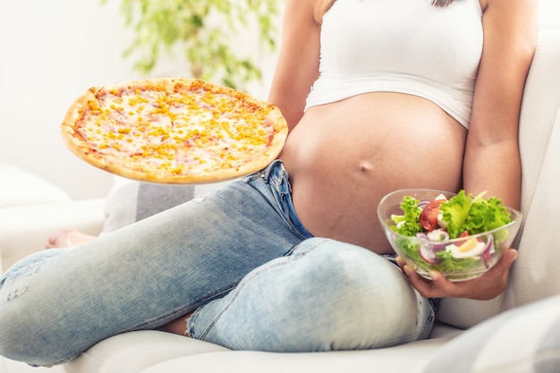 Pregnant girl sitting on a sofa holding salad bowl in one hand and pizza in the other one.