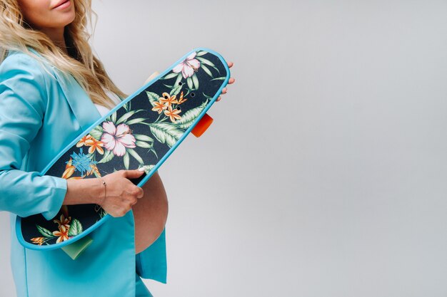 Photo a pregnant girl in turquoise clothes with a skateboard in her hands on a gray background