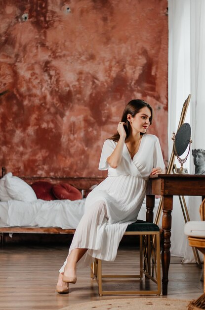 a pregnant girl in a white dress sits at the table and looks in the mirror