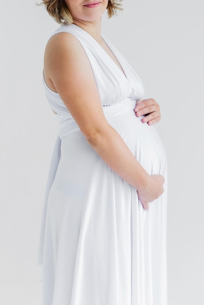 Pregnant girl in a white silk dress hugs her belly and poses on a white background